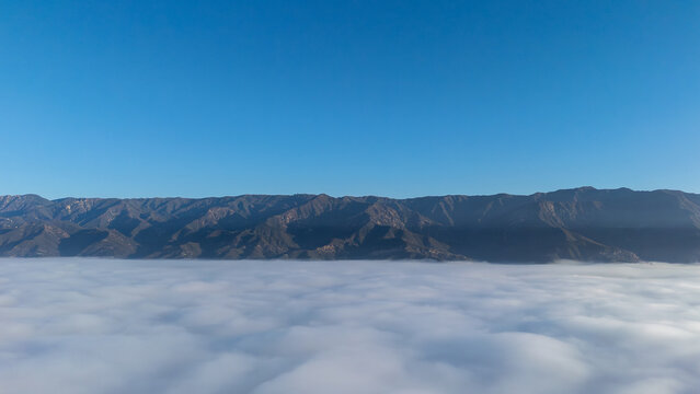 Aerial view of a sea of clouds stretching to meet the base of rugged mountains under a clear blue sky, Montecito, California, United States.