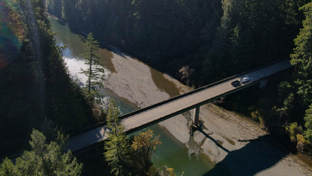 Aerial view of a bridge stretching across a wide, shallow riverbed, flanked by dense green forests under a bright sky, Miranda, California, United States.