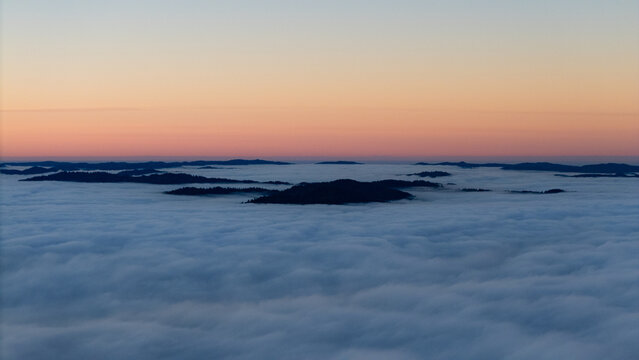Aerial view of dark land formations emerging from a sea of white clouds under a gradient sky painted with soft hues of orange and pink., Miranda, California, United States.