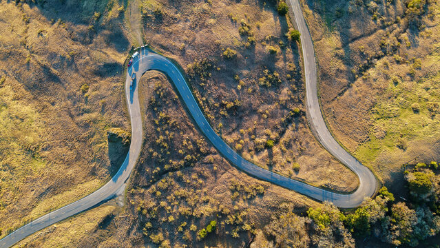 Aerial view of a winding asphalt road slicing through golden, grassy hills, bathed in the warm glow of sunlight, Miranda, California, United States.