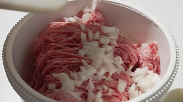 Diced onion pieces fall onto raw minced meat in a mixing bowl. Ingredient prep step before seasoning and shaping patties or meatballs.