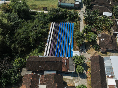 Aerial view of blue and purple textiles laid out to dry under the sun, surrounded by lush trees and traditional Javanese architecture, Surakarta, Central Java, Indonesia.
