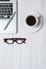 Top view vertical of laptop, coffee cup, green pen and eyeglasses on light wooden background with ample copy space, clean minimal office aesthetic. Concept of modern workspace and lifestyle