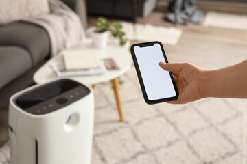 Female hand with mobile phone adjusting air purifier in living room, closeup