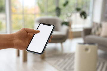 Female hand with mobile phone adjusting air purifier in living room, closeup