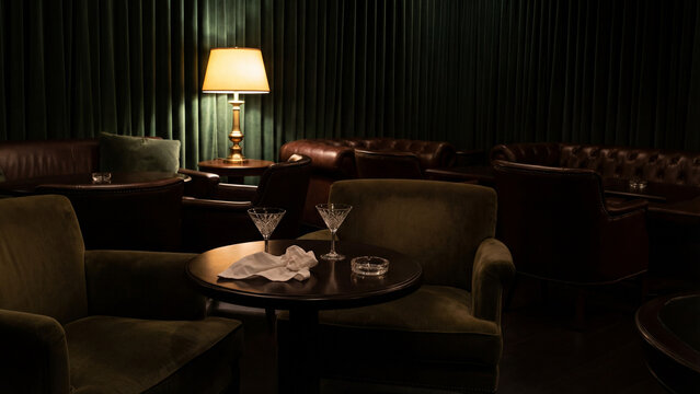 An elegant, dimly lit speakeasy or lounge bar with two martini cocktails on a table. Retro interior with dark green walls, velvet armchairs, and soft lamp light.