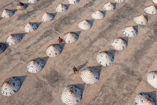 Aerial view of conical hats neatly arranged on a vast, textured surface, with two figures adding a touch of life to the geometric precision, Bhairab, Dhaka Division, Bangladesh.