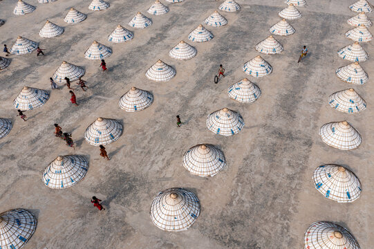 Aerial view of children at play amidst numerous thatched structures casting shadows on the gray ground in Bhairab, Dhaka Division, Bangladesh.