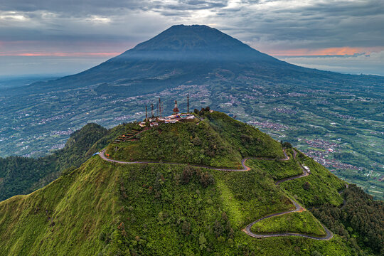 Aerial view of the majestic Telomoyo Mountain, with its lush green slopes and winding roads under a dramatic sky, Magelang, Central Java, Indonesia.