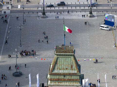Aerial view of the Italian flag waving proudly atop a building overlooking a Piazza Unit&agrave; d'Italia, reflecting the vibrant life below, Trieste, Friuli-Venezia Giulia, Italy.