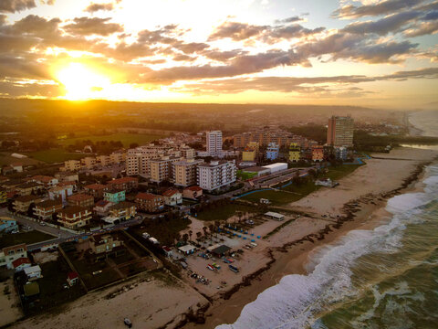 Aerial view of the sun setting behind the buildings along the coast, casting warm hues over the sandy beach and cityscape, Tre Archi, Fermo, Italy.