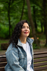 Portrait of confident curly female in nature. Close up portrait of dark haired woman with voluminous curls, smiling confidently against blurred green park background. Ideal for beauty, self love