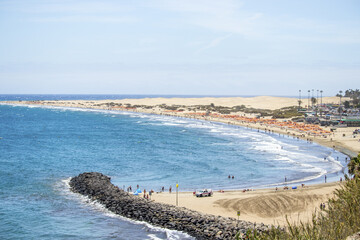 View of turquoise ocean waves crashing against a sandy beach dotted with vibrant orange umbrellas under a clear sky, Gran Canaria, Canary Islands, Spain.