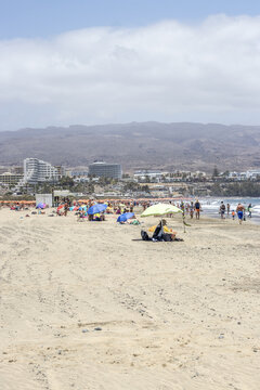 View of a sun-drenched beach where turquoise parasols dot the golden sands, contrasting with the hazy mountains in the distance, Gran Canaria, Canary Islands, Spain.