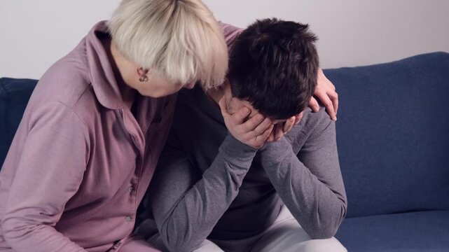 A man sits on the couch, stressed and sad. His wife pities him and provides him with moral support. This is a difficult period in the partners life.