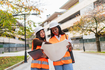 Two women construction workers wearing hard hats and safety vests reviewing building plans outdoors, focused on teamwork and planning