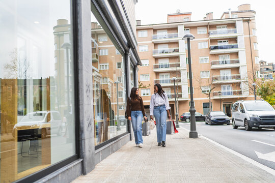 Two diverse women friends walking along city sidewalk carrying shopping bags and smiling, enjoying their time together - Powered by Adobe