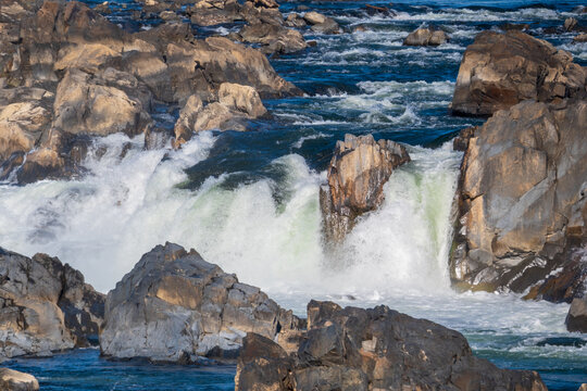 View of powerful, rushing water cascading over rugged, sunlit rocks, creating a dramatic display of nature's force and beauty, Great Falls National Park, Virginia, United States.