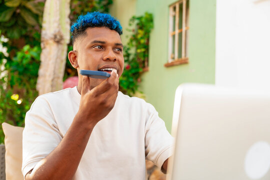 Young latin man with blue hair sending audio message on smartphone while working with laptop outdoors