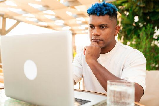 Stylish man with blue hair focused, working remotely on laptop at outdoor cafe, strategizing