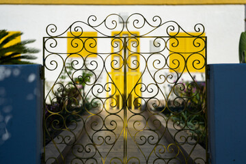 Ornate wrought iron gate with yellow door and shutters in the background