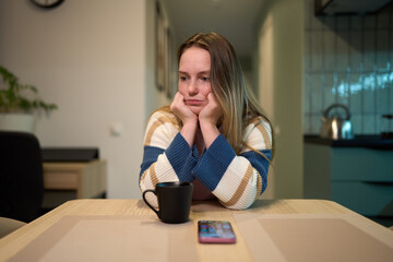 Female resting head on hands at kitchen table with mug and smartphone