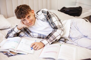 A teenage boy in a plaid shirt lies on his bed and studies with textbooks and notebooks, focusing...