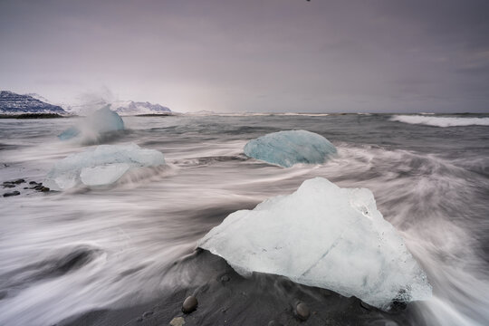 View of glacial ice chunks resting on black sands as waves crash around them under a cloudy sky, Sveitarfelagid Hornafjordur, Sveitarfelagid Hornafjordur, Iceland.