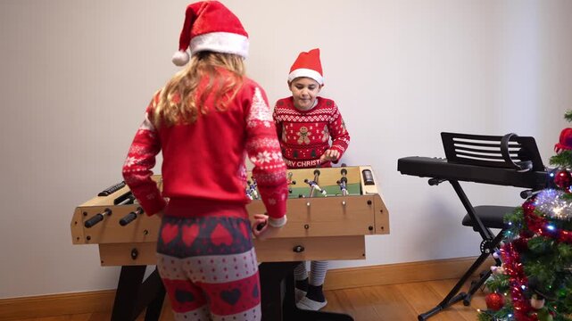 Two happy siblings wearing santa hats and festive sweaters playing with a new foosball table next to a decorated christmas tree