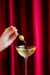 A hand garnishes a cocktail in a martini glass with an olive on a skewer, set against a luxurious red velvet curtain backdrop, evoking a sophisticated, celebratory mood