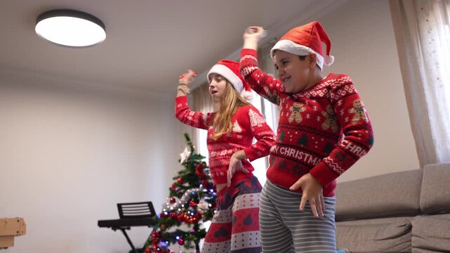 Happy siblings wearing santa hats and festive sweaters playing an active video game at home with a decorated christmas tree in the background - Powered by Adobe