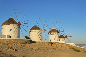 Mykonos, Greece, traditional cylindrical windmills with conical thatched roofs and large wooden blades. The whitewashed structures stand on a stone platform under sunrise light.