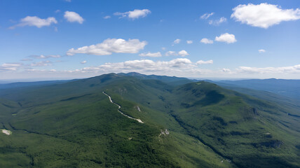 Naklejka premium Aerial view of a lush green mountain range with a winding trail under a clear blue sky and white clouds