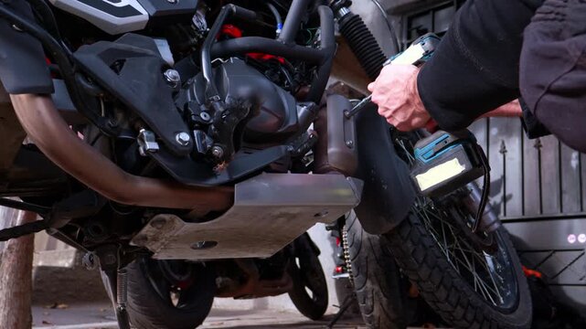 A mechanic repairs a motorcycle outdoors. He's unscrewing a bolt from the muffler resonator cover. He's using a cordless, black-and-blue impact wrench. Left view.