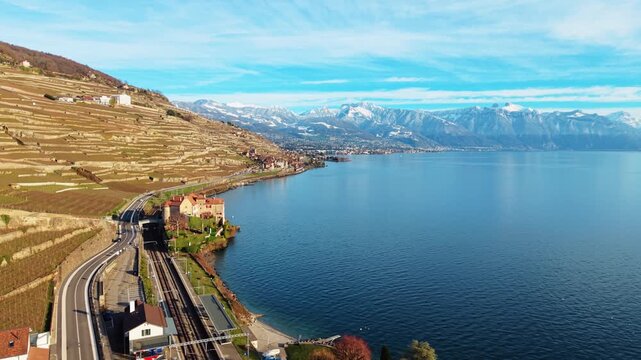 Aerial view of Glerolles Castle nestled by a serene lake, a railway and terraced vineyards with snow-capped mountains in the distance, Saint-Saphorin, Vaud, Switzerland.