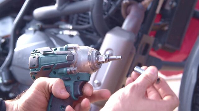 A mechanic repairs a motorcycle outdoors. He installs a bit into a blue and black cordless impact wrench.