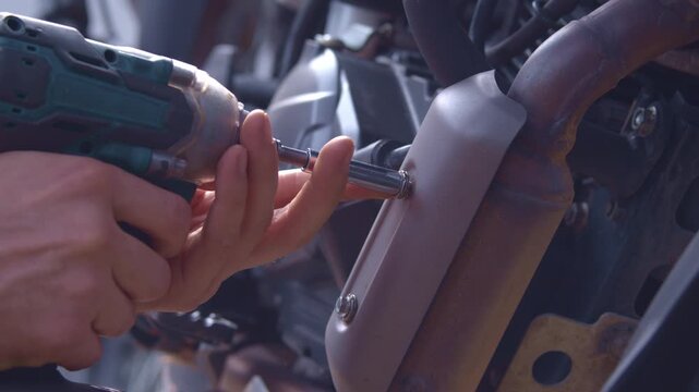 A mechanic repairs a motorcycle outdoors. He's unscrewing a bolt from the muffler resonator cover. He's using a cordless, black-and-blue impact wrench, close-up right top view.