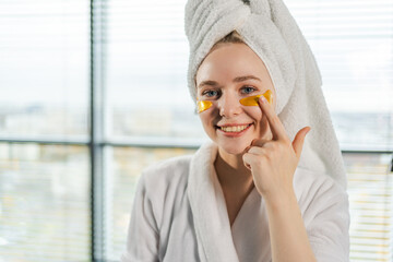 Eye patch beauty treatment. Woman with under eye collagen pads. Girl with patches under eyes towel on head gets facial treatment. Young beautiful woman in robe and towel relaxing in bathroom at home
