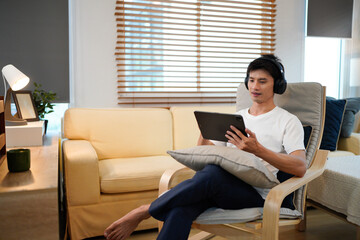 Young Asian man wearing headphones relaxing in cozy living room, smiling while choosing music on tablet