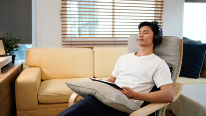 Young Asian man wearing headphones relaxing in cozy living room, smiling while choosing music on tablet