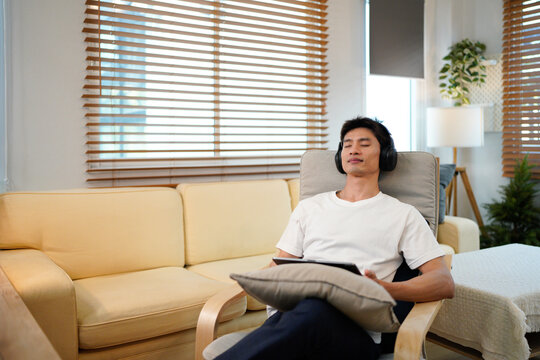 Young Asian man wearing headphones relaxing in cozy living room, smiling while choosing music on tablet - Powered by Adobe