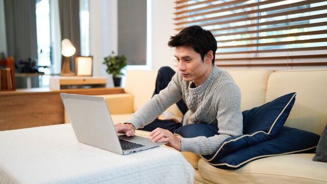 Confident Asian man using laptop while lay down on cozy sofa at modern home - Powered by Adobe