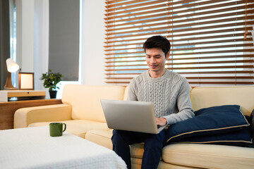 Fototapeta premium Young asian male tech user relaxing holding laptop computer and looking at the screen in living room, Remote Job or work from home concept.