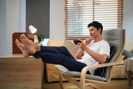 Relaxed young asian man using a smartphone while sitting comfortably in an armchair at living room