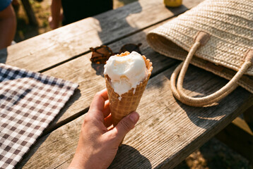 Hand holding a melting vanilla ice cream cone at an outdoor picnic table in summer sunlight, casual leisure and seasonal treat with rustic wood texture and holiday travel vibe