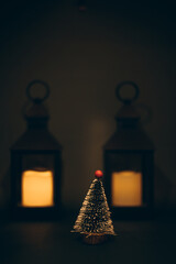 A small decorative Christmas tree with a red topper stands illuminated by warm candle lanterns in a dark, cozy setting.