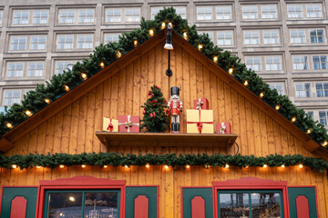Decorated wooden market cabin facade with garlands, lights, gift boxes and a nutcracker figure. Urban office building windows form a patterned background.
