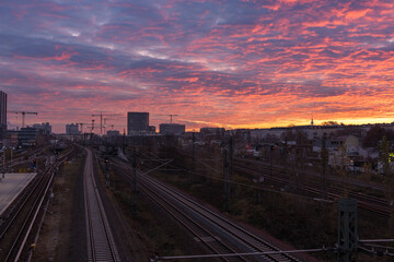 Railway tracks curve into the distance under a vivid red-orange dawn sky. Urban skyline and construction cranes frame a moody Berlin morning.