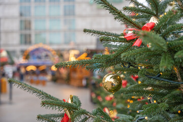 Close-up of a Christmas tree branch with a gold bauble, red ribbon and fairy lights. Soft bokeh background shows a festive winter market in the city.