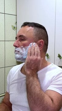 white man shaving with brush and shaving foam looking in the mirror. A man shaves his face in a bathroom with butterflies on the wall and light green tiles.
